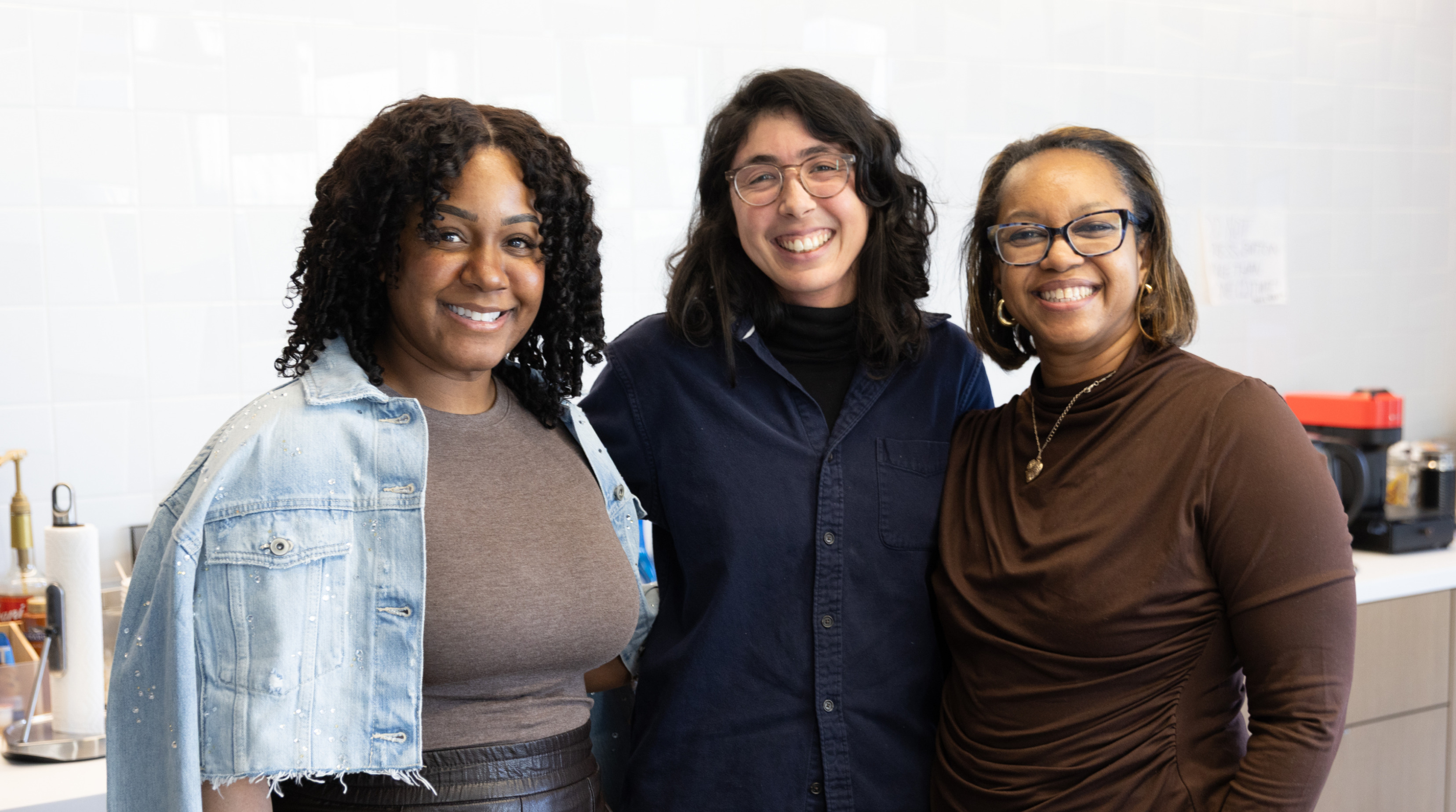 Three smiling colleagues posing together in an office kitchen.
